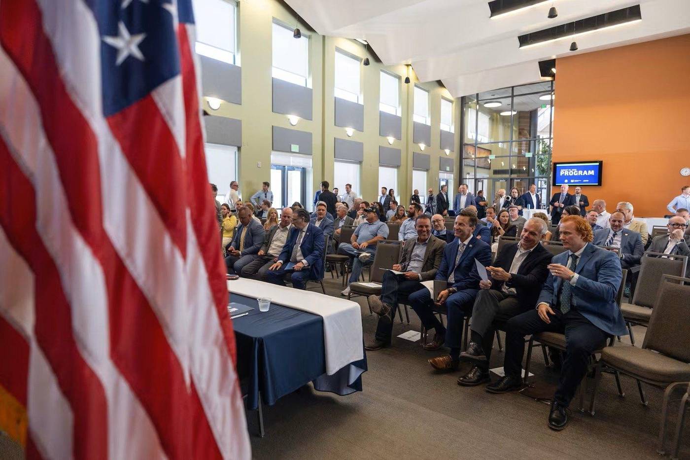 Business and government leaders seated at a public program event, with an American flag in the foreground